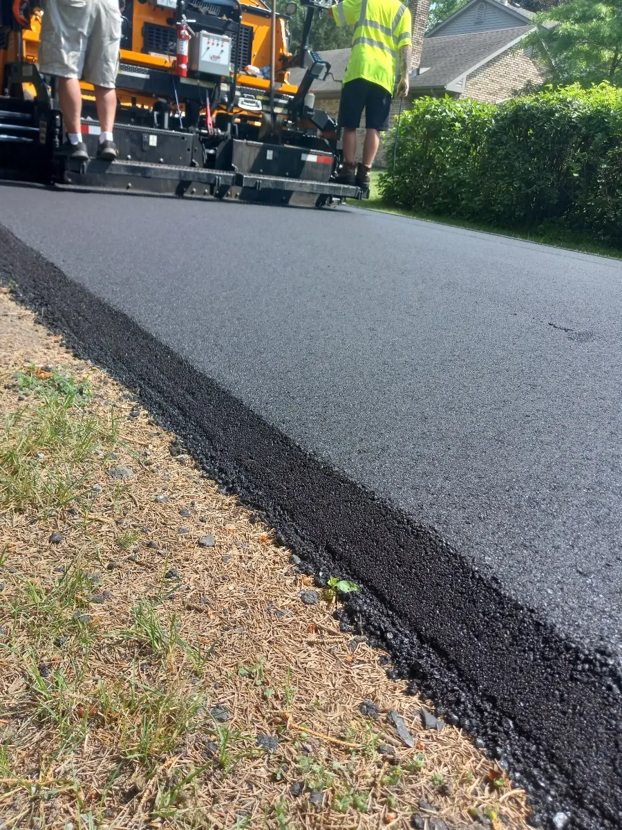 Close-up of a thick, cleanly compacted asphalt edge beside the grass with the paver behind