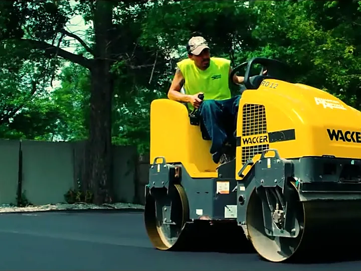 Operator in a safety vest driving a double-drum roller across fresh asphalt
