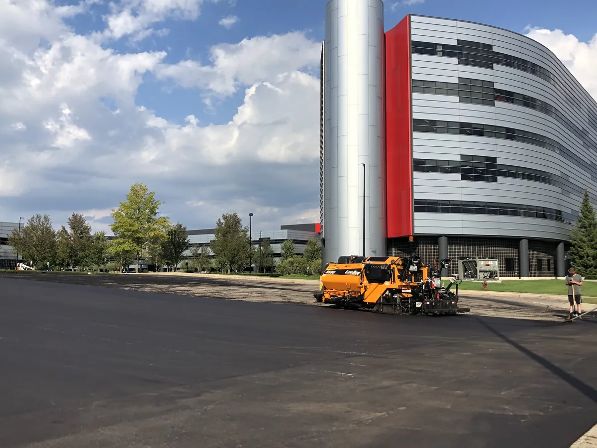 Paver and crew laying a large commercial parking lot in front of a modern office building