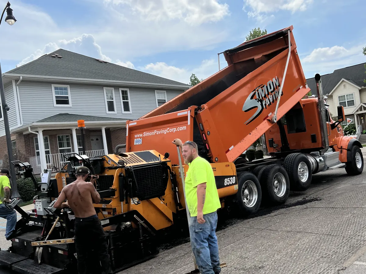 Orange Simon Paving dump truck and paver on a residential street lined with homes